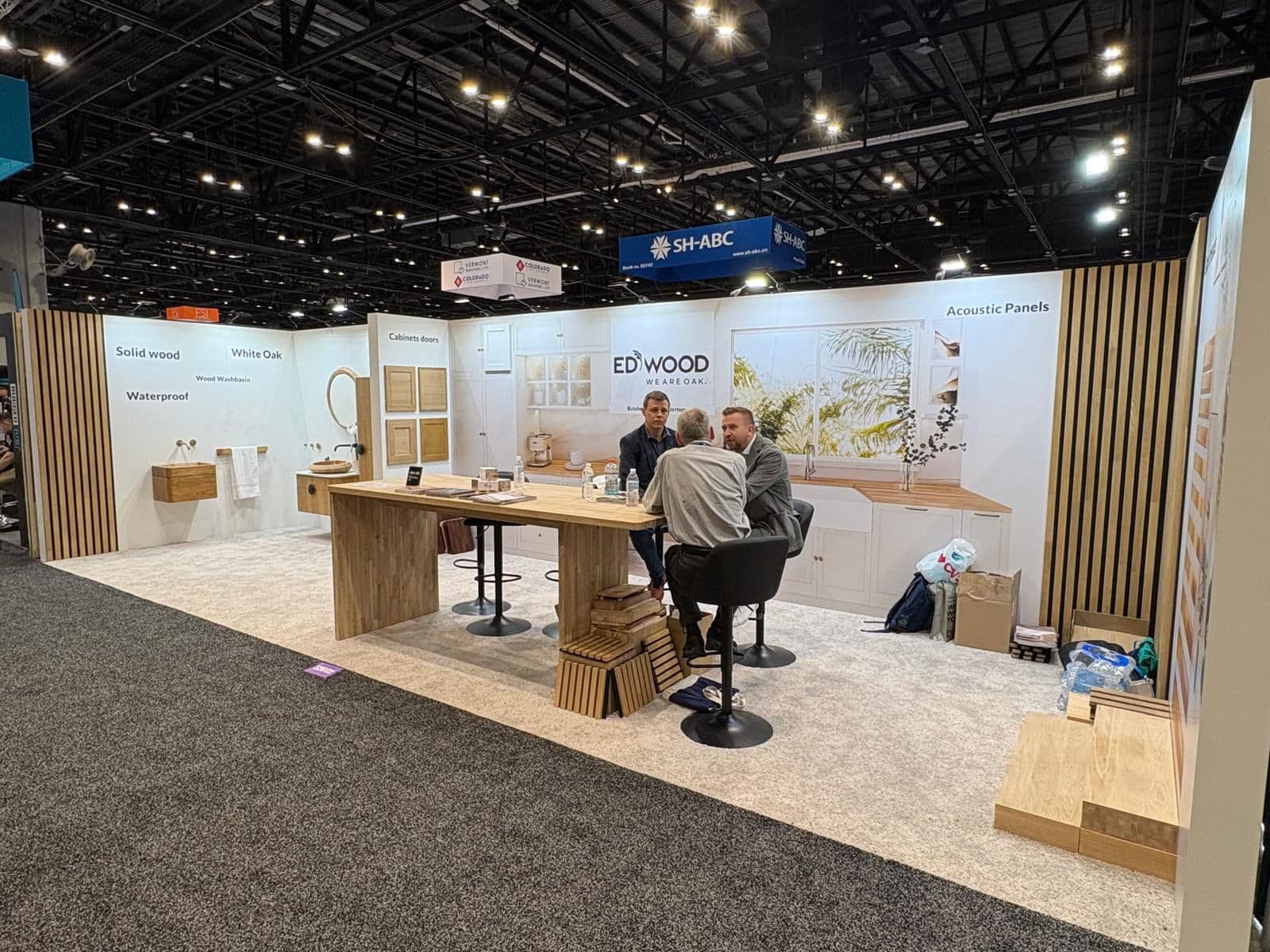 Three men discuss wood products at an Edwood exhibition booth featuring oak kitchen displays.