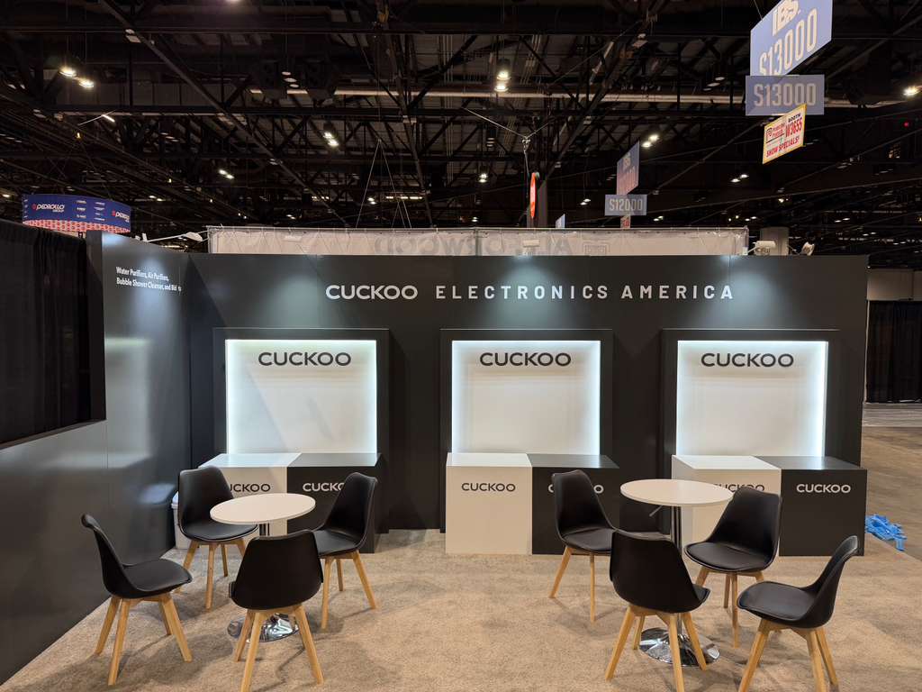 Cuckoo Electronics America trade show booth with black walls, white backlit displays, and seating.