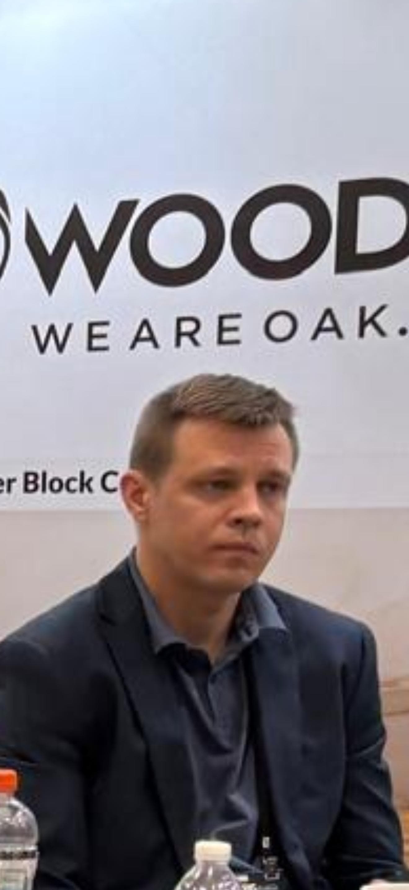 Man in a navy blazer sits in front of a 'WOOD WE ARE OAK' banner.